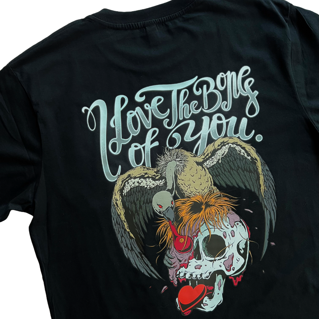 closeup of the back of the black unisex t-shirt. 'I Love The Bones Of You.' is written on the top in a cursive style text. A vulture is perched on top of a decaying skull and eating the remains. A love heart is coming from the skull's mouth