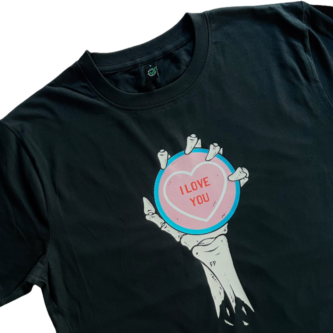 closeup of a black unisex t-shirt with a skeleton hand and forearm holding a oversized love heart candy. I Love You is written on the candy. the colours of the candy are the colours of the transgender flag